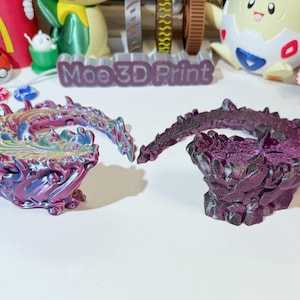 Puede incluir: Dos bases de dragón en miniatura impresas en 3D, una con un acabado morado e iridiscente y la otra con un acabado morado oscuro. Las bases tienen forma de cola de dragón y tienen una superficie texturizada.