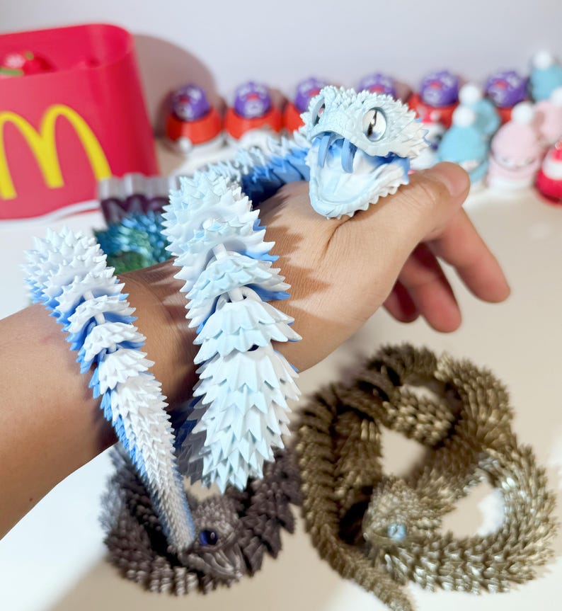 May include: A blue and white snake-shaped toy with a spiky texture. The toy is made of soft, flexible material and is perfect for fidgeting or sensory play. The toy is shown on a person's wrist.