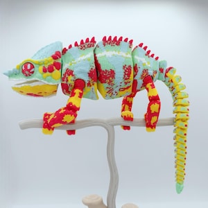 Può includere: Un giocattolo camaleonte articolato e colorato, appollaiato su un ramo di colore chiaro. Il camaleonte presenta un vivace mix di tonalità rosse, gialle, blu e verdi. Il suo corpo e la coda segmentati consentono di posarlo. Il giocattolo è progettato con un aspetto dettagliato e strutturato.
