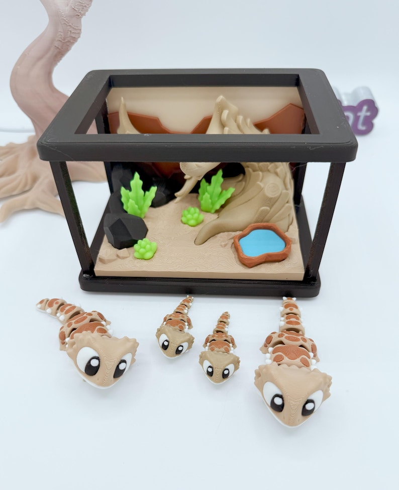Pu&ograve; includere: Un terrario in miniatura con cornice nera, raffigurante un paesaggio desertico con piante verdi, uno stagno blu e sabbia beige. Quattro piccole statuette di geco, marroni e beige, sono disposte davanti al terrario.