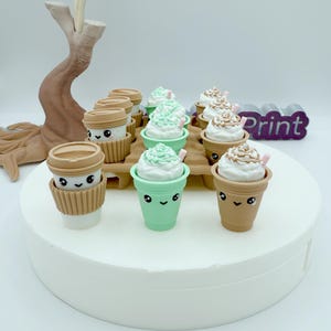 Könnte beinhalten: Eine Sammlung von Miniatur-Kaffee- und Milchshake-Bechern im Cartoon-Stil. Die Becher sind in verschiedenen Farben, darunter Braun und Mintgrün, mit Schlagsahne-Toppings und rosa Strohhalmen. Die Becher haben lächelnde Gesichter.