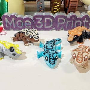 以下が含まれることがあります： 異なる色とパターンで3Dプリントされた5つのヤモリの置物。ヤモリは尻尾を丸めて歩いているポーズをとっています。ヤモリは青、黄、オレンジ、茶色、白でプリントされています。