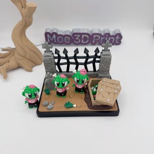 May include: A miniature graveyard scene featuring green zombie figures with pink brains and black outfits. The scene includes a tombstone, a coffin, and a black fence. The text "Moe 3D Print" is visible in the background.