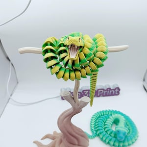 May include: A 3D-printed articulated snake in shades of green and yellow, posed with its mouth open. The snake is mounted on a brown tree-like stand, with a second coiled snake in blue and green at the base.