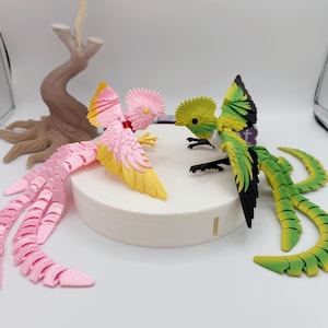 May include: Two colorful bird figurines. One is pink and yellow, the other is green and yellow. They are positioned on a white platform. A brown tree-like sculpture is in the background.