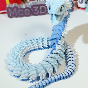 May include: A 3D printed blue and white snake figurine with a spiky texture. The snake is curled up in a circle with its head raised. The figurine is made of plastic and is about 10 inches long.