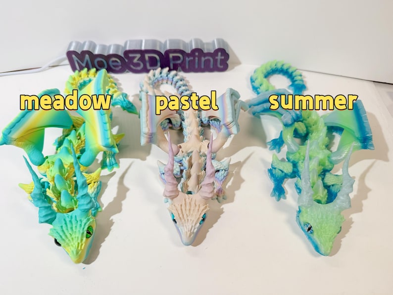 May include: Three 3D printed dragon figurines in different color schemes. The first dragon is green and yellow with the word "meadow" in front of it. The second dragon is white and blue with the word "pastel" in front of it. The third dragon is green and blue with the word "summer" in front of it.