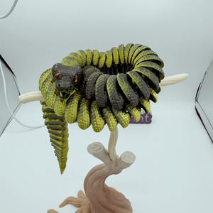 Op de afbeelding: Gedetailleerde sculptuur van een opgerolde slang, voornamelijk groen en zwart, met rode ogen. De slang zit op een gestileerde tak, met een bruine basis en cr&egrave;mekleurige takken. De sculptuur staat tegen een witte achtergrond.