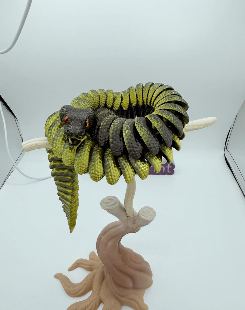 Op de afbeelding: Gedetailleerde sculptuur van een opgerolde slang, voornamelijk groen en zwart, met rode ogen. De slang zit op een gestileerde tak, met een bruine basis en cr&egrave;mekleurige takken. De sculptuur staat tegen een witte achtergrond.
