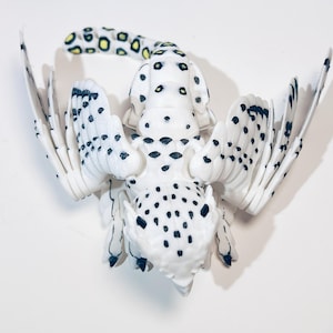May include: A white and black spotted owl figurine with outstretched wings. The owl has yellow accents on its tail feathers.