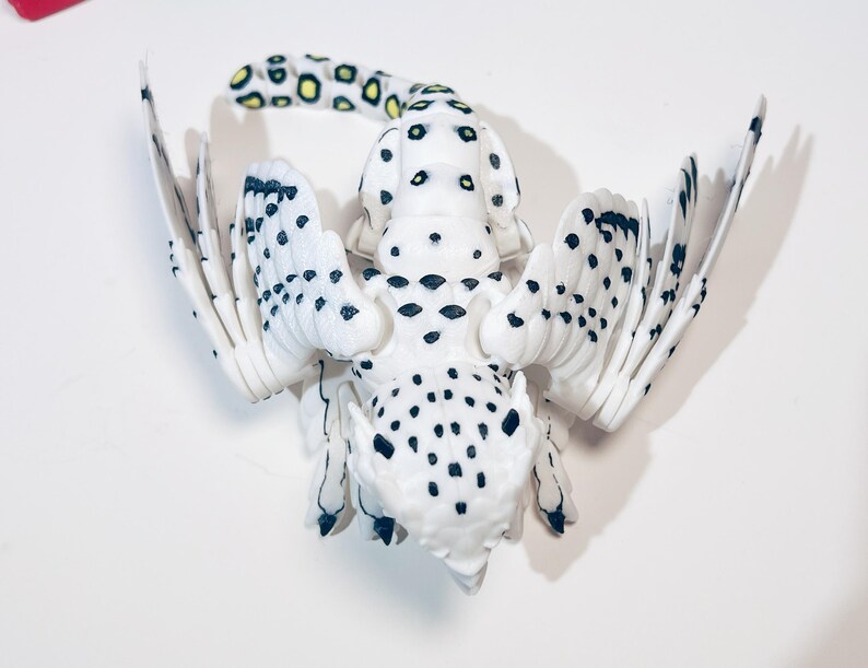 May include: A white and black spotted owl figurine with outstretched wings. The owl has yellow accents on its tail feathers.