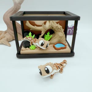 Pu&ograve; includere: Un terrario in miniatura con un geco dei cartoni animati e una scena desertica. Il contenitore con cornice nera presenta una base marrone, un albero e un piccolo stagno blu. Un secondo geco &egrave; posizionato fuori dal terrario. I gechi sono marroni con macchie marroni.
