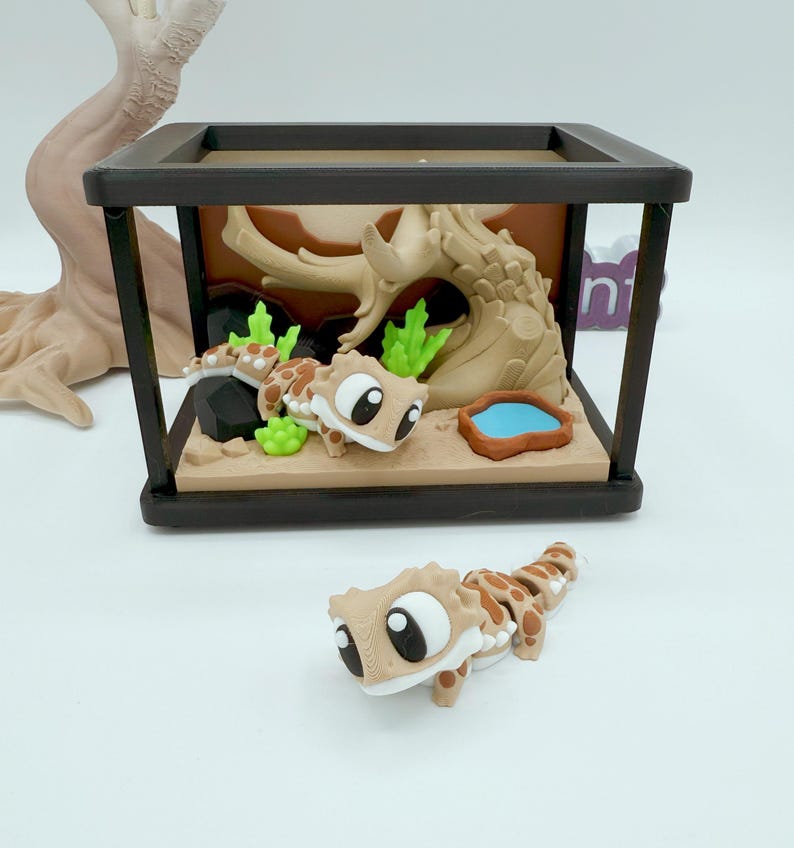 Pu&ograve; includere: Un terrario in miniatura con un geco dei cartoni animati e una scena desertica. Il contenitore con cornice nera presenta una base marrone, un albero e un piccolo stagno blu. Un secondo geco &egrave; posizionato fuori dal terrario. I gechi sono marroni con macchie marroni.