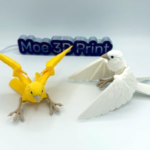 以下が含まれることがあります： 3Dプリントされた鳥の置物2つ。片方は黄色で翼を広げ、もう片方は白で翼を広げています。鳥は白い面にいます。「Moe 3D Print」の文字が背景に見えます。