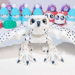 May include: A white and black articulated owl toy with large, black eyes. The owl has its wings spread out and is posed in a standing position.
