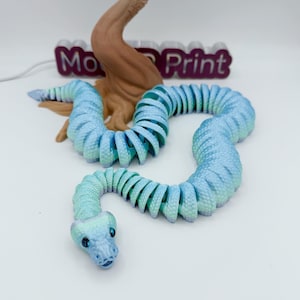 Op de afbeelding: Een 3D-geprint, beweeglijk slangen speelgoed in tinten blauw en groen. De slang is opgerold en heeft een gedetailleerde kop met zichtbare ogen. De achtergrond bevat een bruine boomachtige structuur en de tekst "Mo Print".