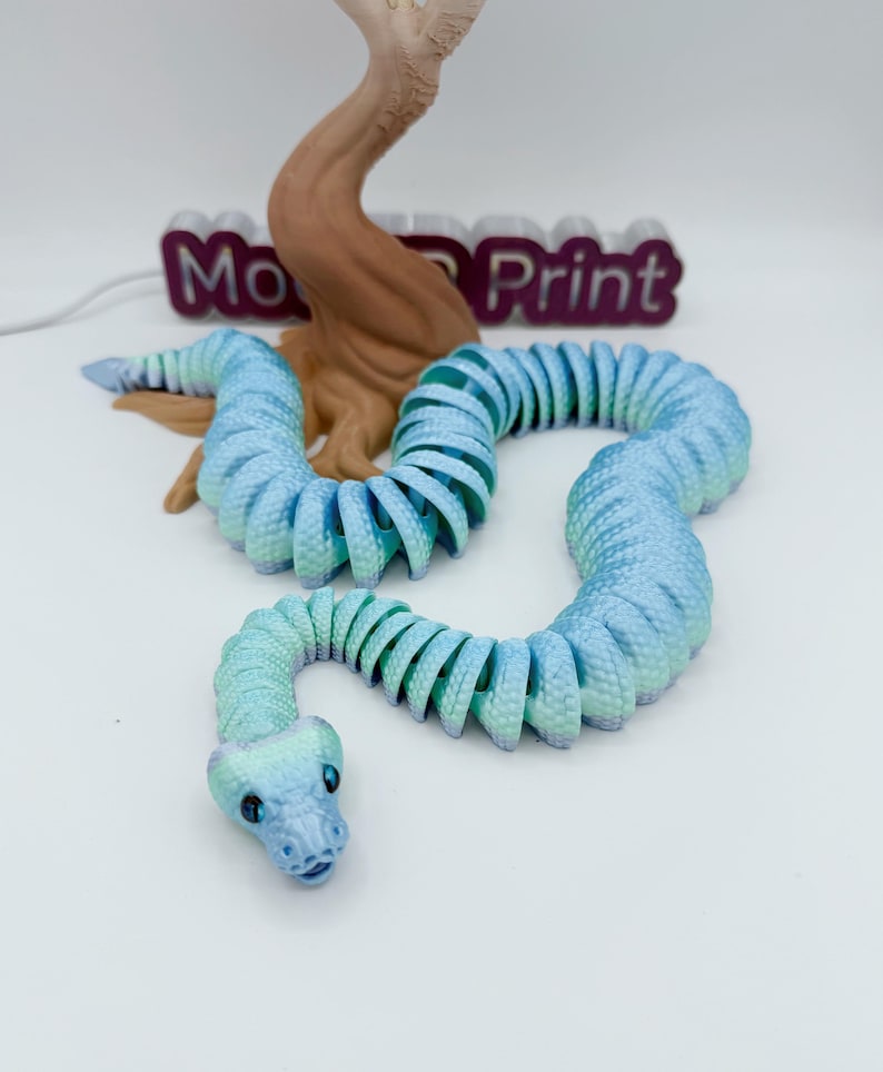 Op de afbeelding: Een 3D-geprint, beweeglijk slangen speelgoed in tinten blauw en groen. De slang is opgerold en heeft een gedetailleerde kop met zichtbare ogen. De achtergrond bevat een bruine boomachtige structuur en de tekst "Mo Print".