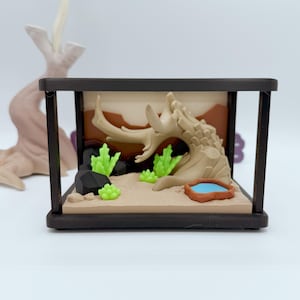 Pu&ograve; includere: Diorama di terrario in miniatura con un paesaggio desertico con un tronco d'albero marrone chiaro, piante verdi e un piccolo stagno blu. La scena &egrave; racchiusa in una cornice rettangolare marrone scuro.