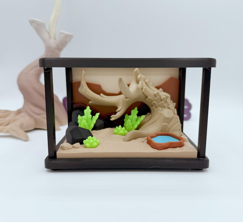 Pu&ograve; includere: Diorama di terrario in miniatura con un paesaggio desertico con un tronco d'albero marrone chiaro, piante verdi e un piccolo stagno blu. La scena &egrave; racchiusa in una cornice rettangolare marrone scuro.