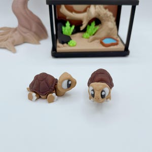 Pu&ograve; includere: Due piccole figurine di tartarughe in stile cartone animato con gusci marroni e corpi beige. Una tartaruga &egrave; in posizione di gattonamento, l'altra &egrave; in posizione eretta. Un terrario in miniatura con un paesaggio e un albero sullo sfondo.