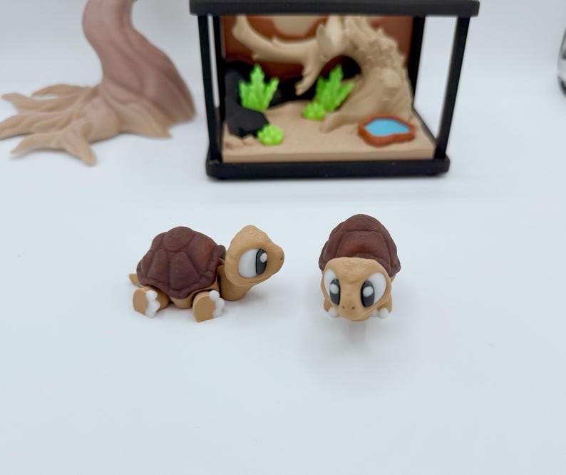 Pu&ograve; includere: Due piccole figurine di tartarughe in stile cartone animato con gusci marroni e corpi beige. Una tartaruga &egrave; in posizione di gattonamento, l'altra &egrave; in posizione eretta. Un terrario in miniatura con un paesaggio e un albero sullo sfondo.