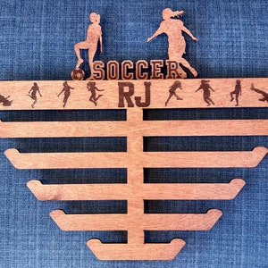 Pu&ograve; includere: Porta medaglie in legno con sei ganci per appendere le medaglie. Il porta medaglie &egrave; decorato con un tema calcistico, tra cui un pallone da calcio, due calciatrici e il testo "SOCCER RJ".