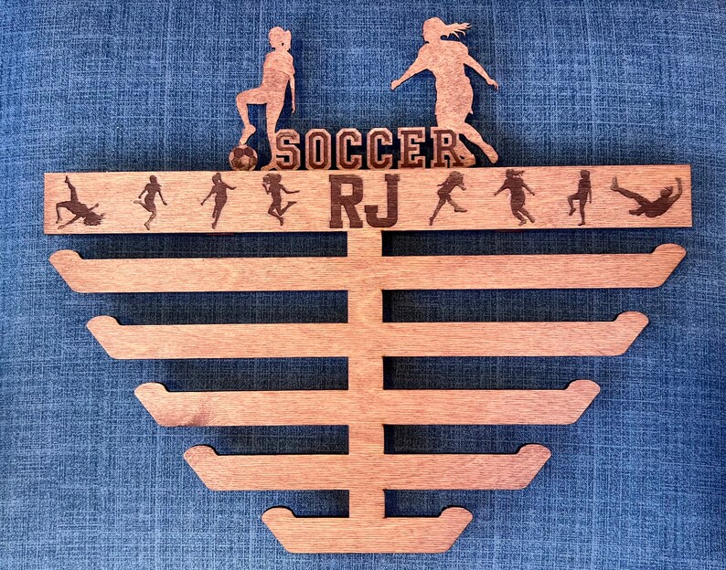 Pu&ograve; includere: Porta medaglie in legno con sei ganci per appendere le medaglie. Il porta medaglie &egrave; decorato con un tema calcistico, tra cui un pallone da calcio, due calciatrici e il testo "SOCCER RJ".