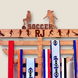 Pu&ograve; includere: Un porta medaglie in legno con le parole "SOCCER RJ" e una silhouette di un calciatore. Il porta medaglie ha cinque file per le medaglie ed &egrave; decorato con una macchia marrone.