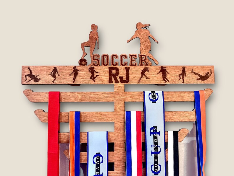 Pu&ograve; includere: Un porta medaglie in legno con le parole "SOCCER RJ" e una silhouette di un calciatore. Il porta medaglie ha cinque file per le medaglie ed &egrave; decorato con una macchia marrone.