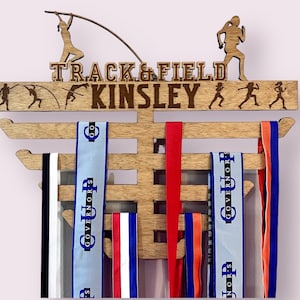 May include: Wooden medal display rack with the words "TRACK & FIELD" and "KINSLEY" engraved on the top. The rack has 8 hooks for hanging medals and ribbons. The ribbons are blue, red, white, and black.