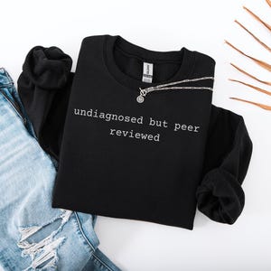 Pode incluir: Camisola de moletom preta com o texto "undisagnosed but peer reviewed" em letras brancas.