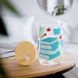 Könnte beinhalten: Ein durchsichtiger Glasbecher in Dosenform mit Bambusdeckel und Strohhalm. Der Becher ist mit einem Muster aus blauen Büchern, roten Äpfeln und Bleistiften verziert. Der Deckel ist rund und aus hellem Holz.