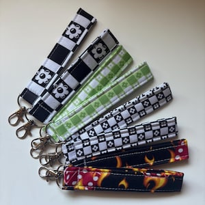 Op de afbeelding: Een verzameling stoffen polsbandjes met zilverkleurige draaisluitingen. De bandjes hebben verschillende patronen, waaronder zwart-wit dambord met bloemmotieven, groene bloemen en een zwarte achtergrond met vlammen en dobbelstenen.