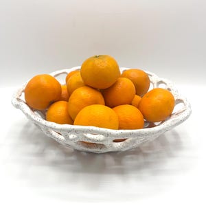 May include: A white ceramic fruit bowl filled with bright orange tangerines. The bowl has a textured, openwork design. The tangerines are round and have a vibrant orange color, suggesting freshness. The arrangement is on a white background.