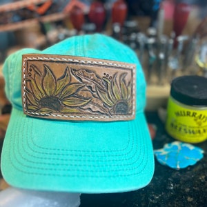 Puede incluir: Una gorra de béisbol azul turquesa con un parche de cuero que presenta un diseño tallado de dos girasoles. El parche está cosido alrededor de los bordes con hilo blanco.