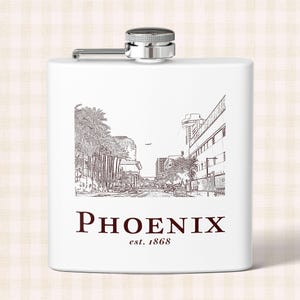 May include: A white metal flask with a silver screw-top cap. The front features a dark brown line-art illustration of a Phoenix city street scene with palm trees and buildings, with the text PHOENIX and est. 1868 printed below the image.