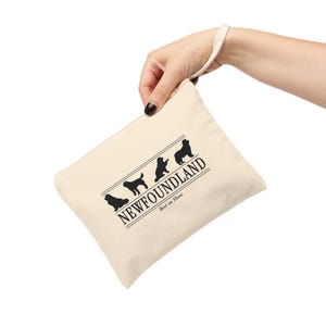 May include: A cream-colored canvas zipper pouch with a wrist strap. The front features a black graphic of three Newfoundland dog silhouettes above the text NEWFOUNDLAND and the smaller text Best in Show, framed by two horizontal lines.