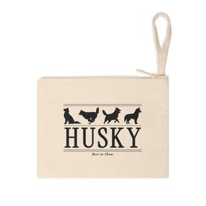 May include: A beige canvas zipper pouch with a wrist strap. The front features a black graphic of four husky silhouettes above the text HUSKY and the smaller text Best in Show, all enclosed between two sets of horizontal black lines.
