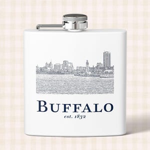 May include: A white rectangular metal flask with a silver screw-top cap. The front features a black line-art illustration of the Buffalo city skyline above the dark blue text "BUFFALO" and "est. 1832" in a serif font centred on the body.