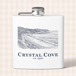 May include: A white metal flask with a silver screw-top cap. The front features a dark blue line-art illustration of a coastal landscape with cliffs and a beach, with the text "CRYSTAL COVE" and "est. 1920" printed in dark blue below the image.