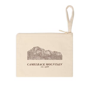 May include: Cream-colored zippered pouch featuring a detailed illustration of Camelback Mountain. The text "CAMELBACK MOUNTAIN est. 1968" is printed below the mountain graphic. The pouch has a loop handle and a gold-colored zipper.