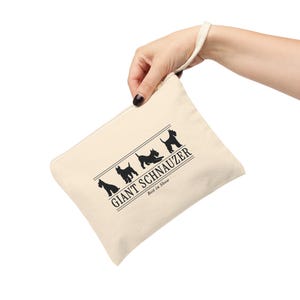 May include: A beige canvas zipper pouch with a wrist strap featuring a black graphic of three Giant Schnauzer dog silhouettes. The text reads GIANT SCHNAUZER and Best in Show, framed by horizontal lines, printed in a clean, dark serif font.