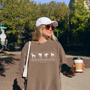 Può includere: Una felpa marrone a girocollo con una grafica bianca di quattro sagome di cani Bracco Tedesco, il testo "GERMAN SHORTHAIRED POINTER" e "Best in Show", un berretto bianco, una borsa a tracolla bianca e un bicchiere di caffè usa e getta.