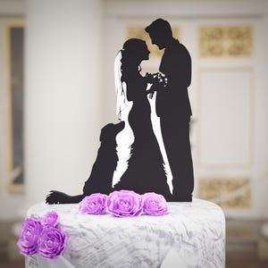 Puede incluir: Adorno para tarta de silueta negra que representa a una novia y un novio, con un perro. La pareja se toma de la mano y la tarta está decorada con rosas moradas. El adorno se coloca sobre una tarta blanca.