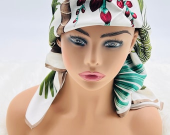 May include: A white silk scarf with a botanical print featuring green leaves, brown stems, and red flower buds. The scarf is draped over a mannequin head, showcasing its design and shape. The edges are finished with a light brown border.