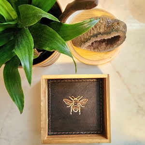 May include: A square, wooden coaster with a dark brown leather insert. A gold-colored bee is centered on the leather. The coaster is surrounded by a light brown wooden frame. The background includes a potted plant and a geode.