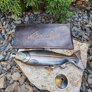 May include: A stainless steel fish-shaped flask with a silver finish, a brown leather wallet with a mountain and tree design, and a silver cap. The flask is lying on a rock.