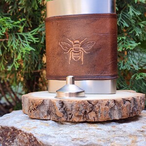 May include: Stainless steel flask with a brown leather wrap featuring an embossed bee design. Includes a silver funnel. The flask is displayed on a wooden slice, with a natural green background.