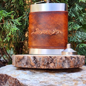 May include: A stainless steel hip flask with a brown leather sleeve, embossed with a mountain range design. The flask is on a wooden slab, with a small funnel. The background is a green, leafy plant.