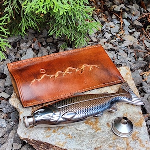 May include: A stainless steel flask shaped like a fish, with a matching funnel, rests on a stone. A brown leather case with a mountain range design is behind the flask. The flask has a silver cap.